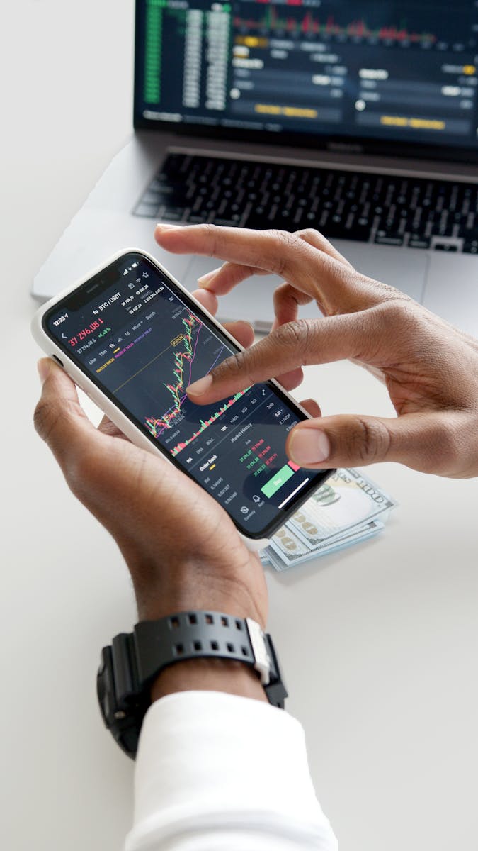 Photo by Anna Nekrashevich Hands using smartphone trading app with charts, dollar bills in background.