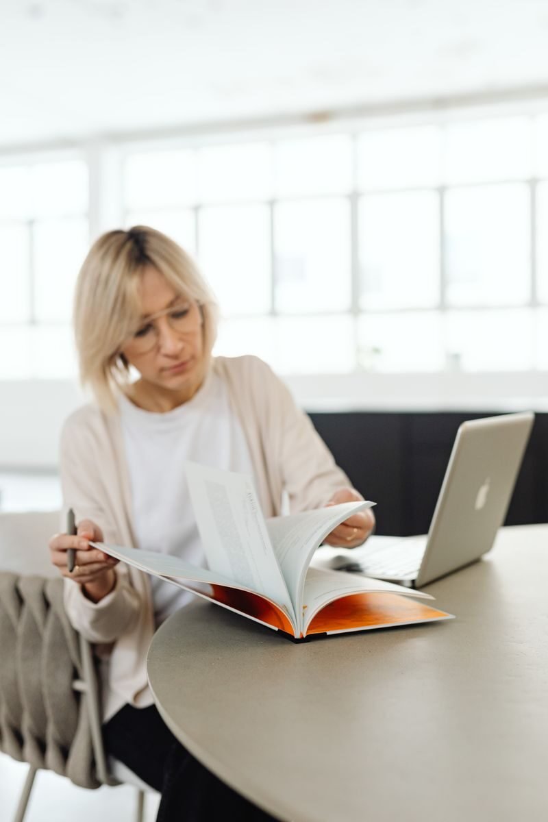 A woman reading a book and working on a laptop in a bright indoor setting.